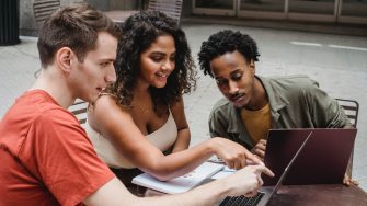 A group of three students sit together in conversation, facing an open laptop.