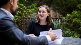 A woman looks inspired while discussing the contents of a book that a man has out on the table where they are seated at an outdoor cafe.